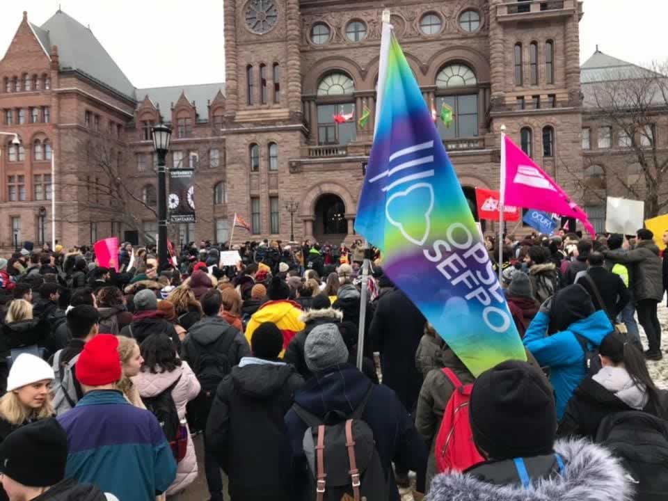 Large group of workers holding flags, including OPSEU flags, during a rally at Queen's Park.