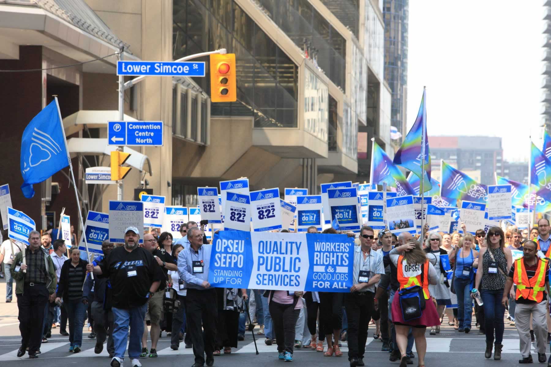 Large group of OPSEU members holding signs, flags, and a "Quality Public Services" banner march in downtown Toronto during Convention 2015