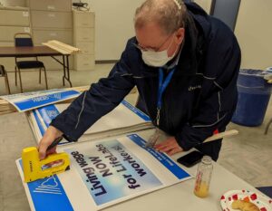 A man learns over a picket sign laid flat on a table as he assembles it