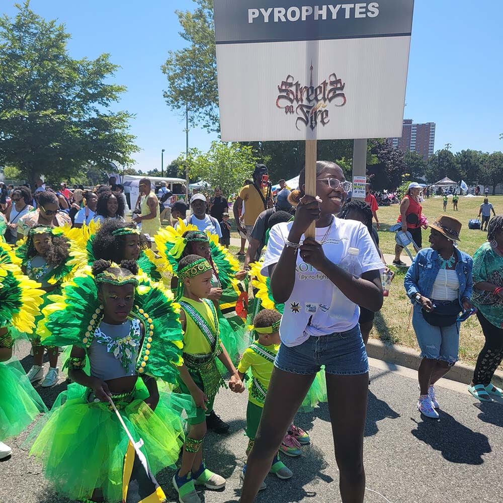Woman with sign reading "Pyrophytes" with a group of children wearing green and yellow for Carnival.