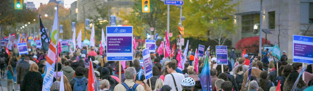 Thousands of people, including in the foreground many carrying OPSEU/SEFPO flags and signs, march towards Queen's Park.