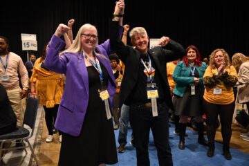 JP Hornick and Laurie Nancekivell stand together at Convention, holding their hands up together in celebration.