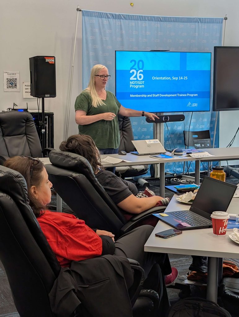 First Vice-President/Treasurer Laurie Brown stands at the head table, addressing several participants seated in chairs. The screen behind Nancekivell says "2026 MDT/SDT Program"