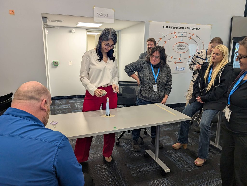 A group of people stand around a table watching one person about to balance a rock on top of a homemade tower. 