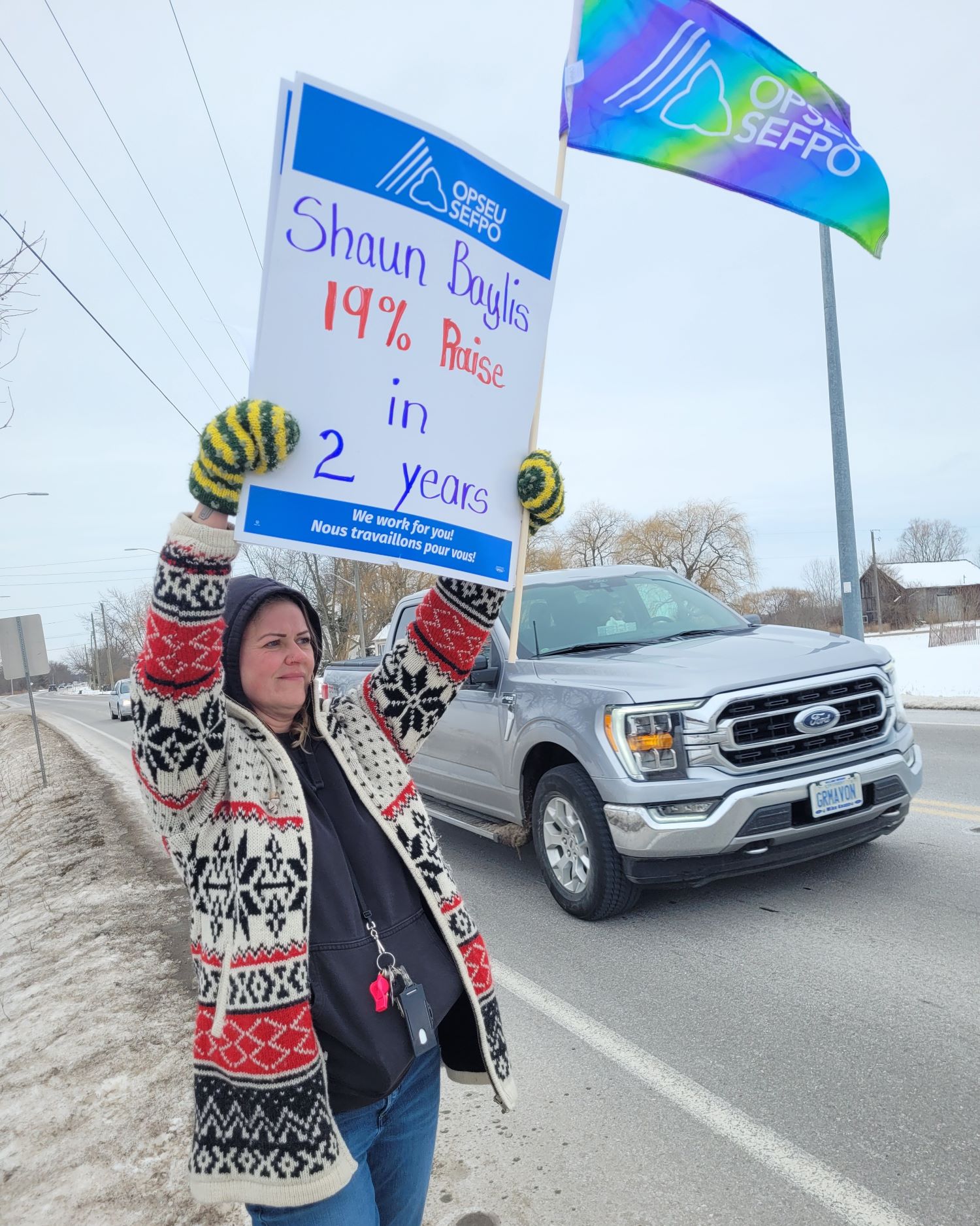 A person standing outside next to a road holding a picket sign that says "Shaun Baylis 19% raise in 2 years"