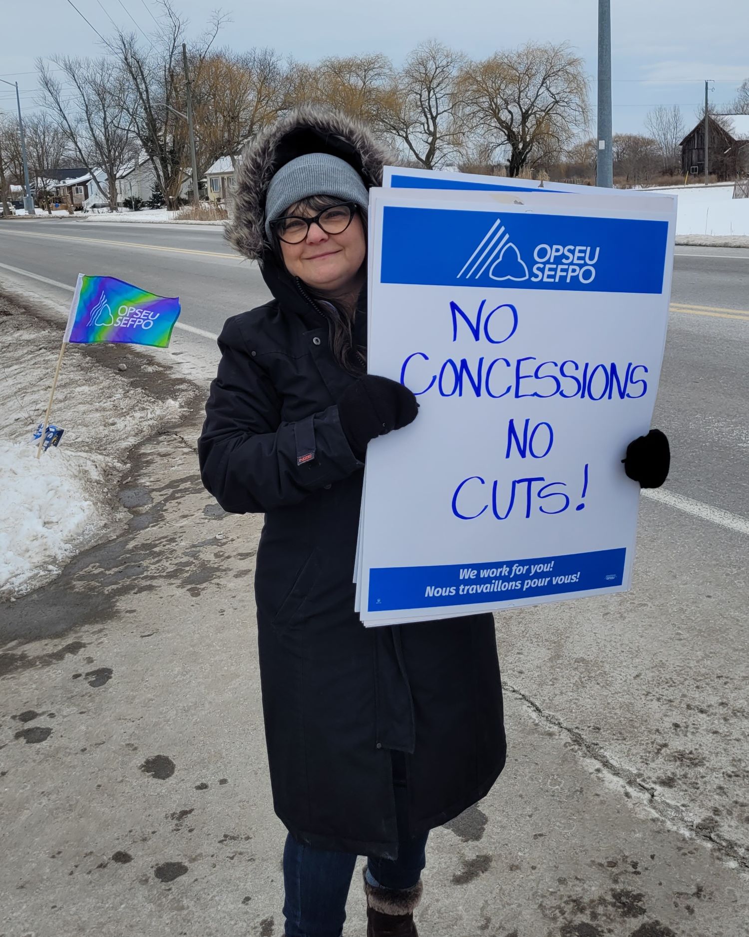 Person outside in winter next to a road holding picket sign that says "No concessions, no cuts!"