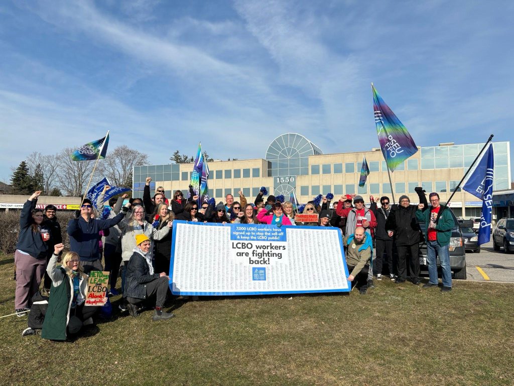 Crowd of people standing together with picket signs, OPSEU/SEFPO flags, and a large banner with thousands of signatures of LCBO workers