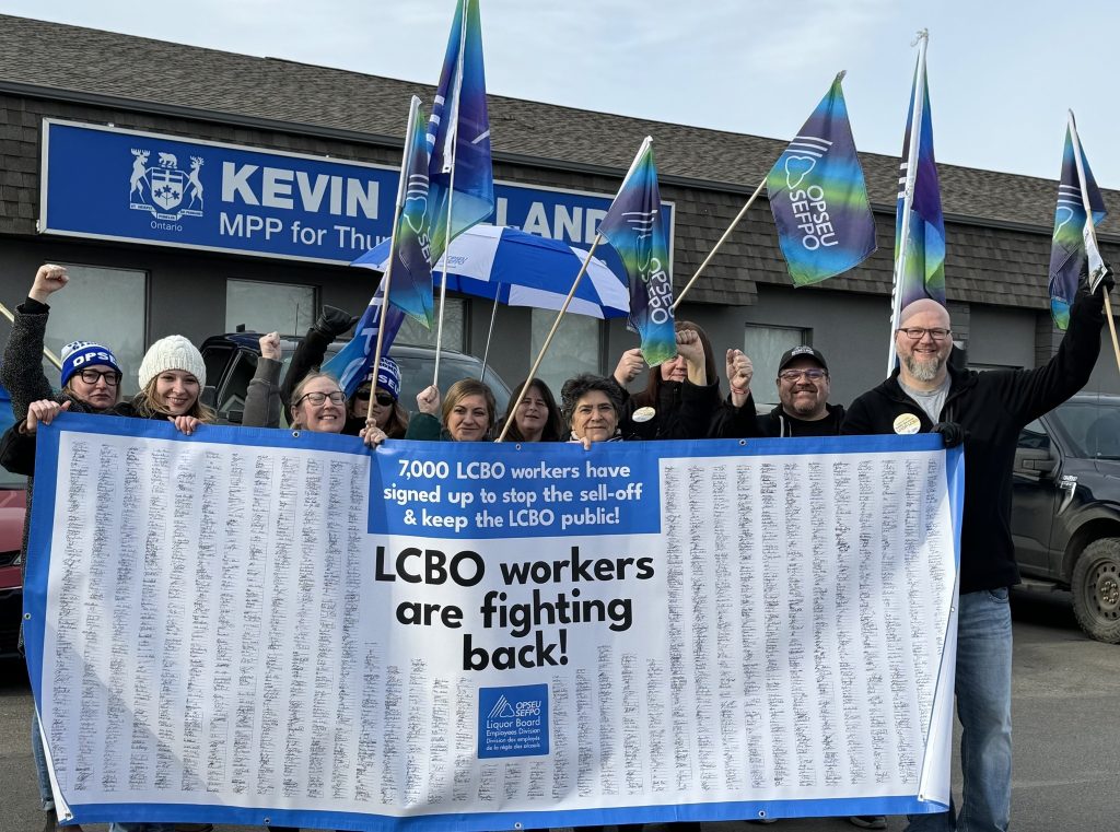 Crowd of people standing together with OPSEU/SEFPO flags, and a large banner with thousands of signatures of LCBO workers