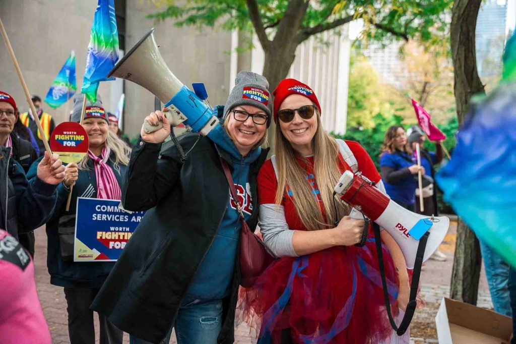 Two smiling people, both holding megaphones, march in a crowd of protesters with OPSEU/SEFPO signs and flags.
