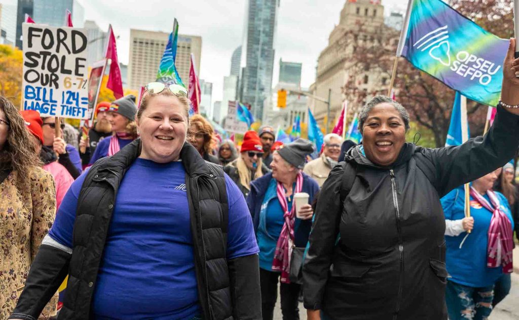Two people, both smiling broadly, march in a crowd of protesters with OPSEU/SEFPO flags and a sign that says "Ford stole. Bill 124 = wage theft"